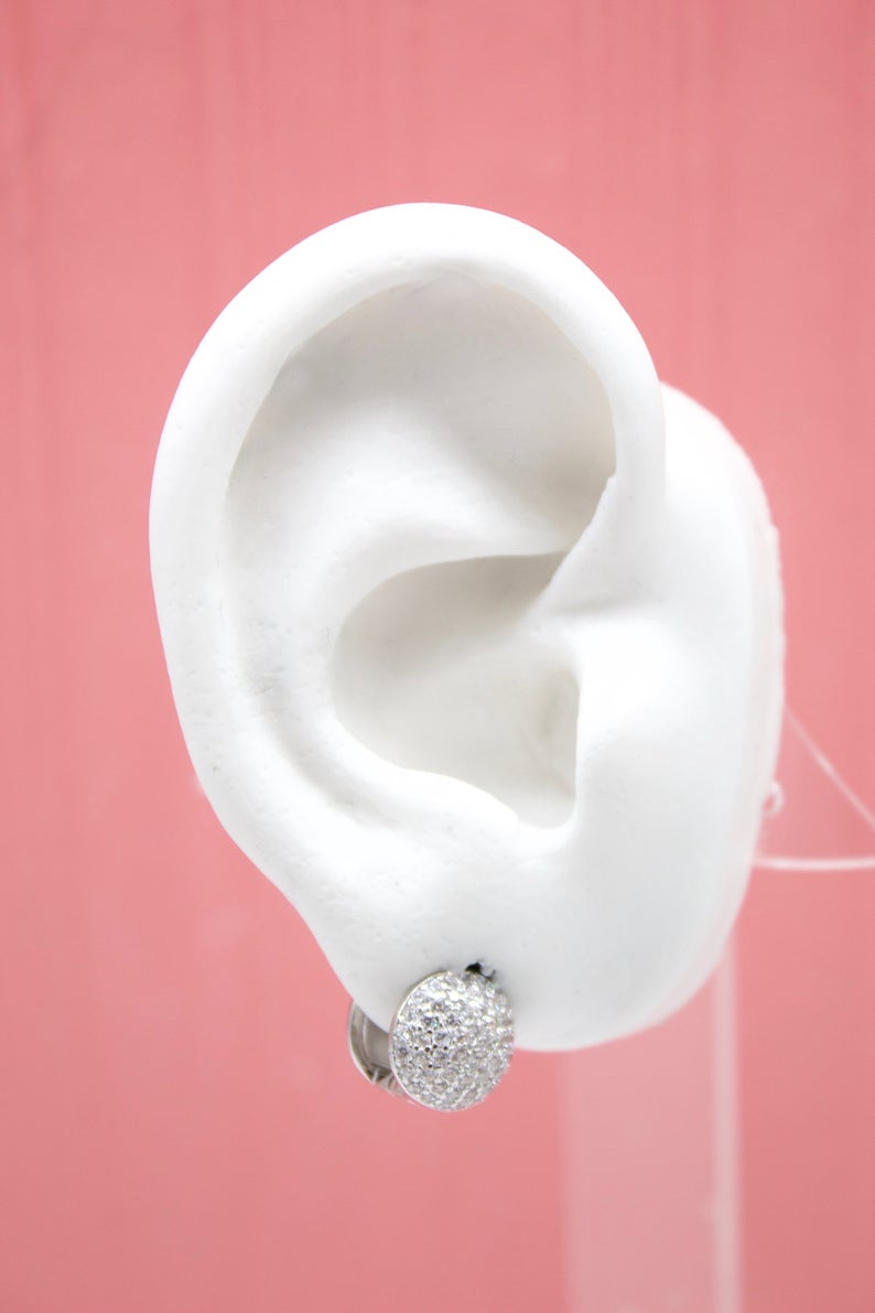 A white model ear showcases a sparkling pave-set diamond huggie hoop earring against a soft pink background.