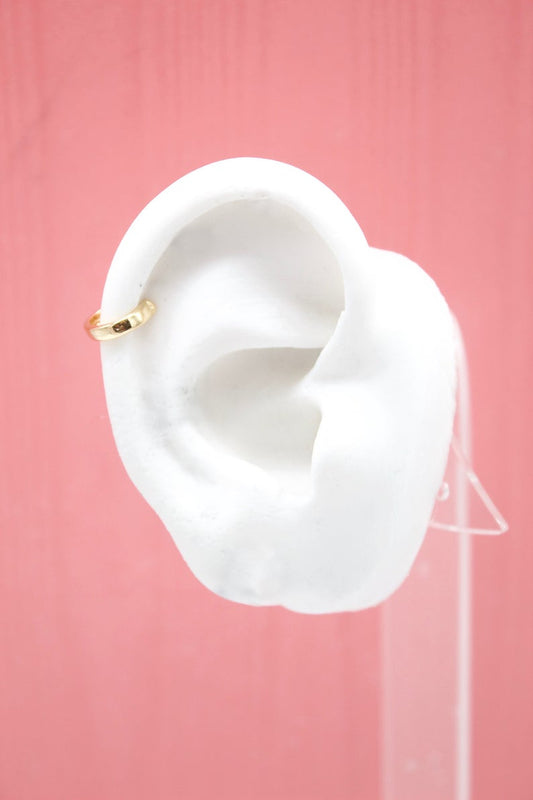 A small gold hoop cartilage earring adorns a white mannequin ear against a pink background.