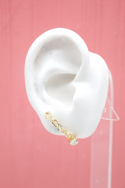 A white mannequin ear displays a gold chain earring featuring interlocking links and a dangling crystal stone against a pink background.