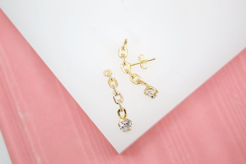 A pair of gold chain earrings dangle on a white surface with a pink background, featuring delicate link drops and sparkling stone accents.