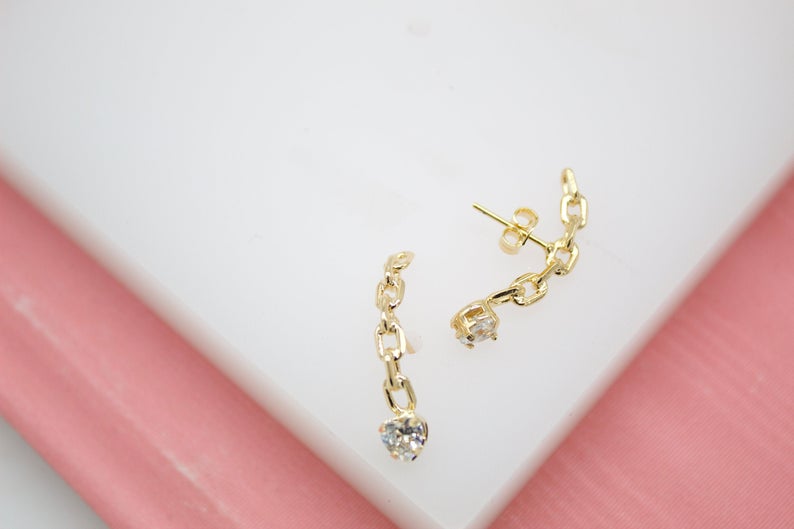A pair of delicate gold chain earrings featuring sparkling zirconia charms rests on a white display surface.