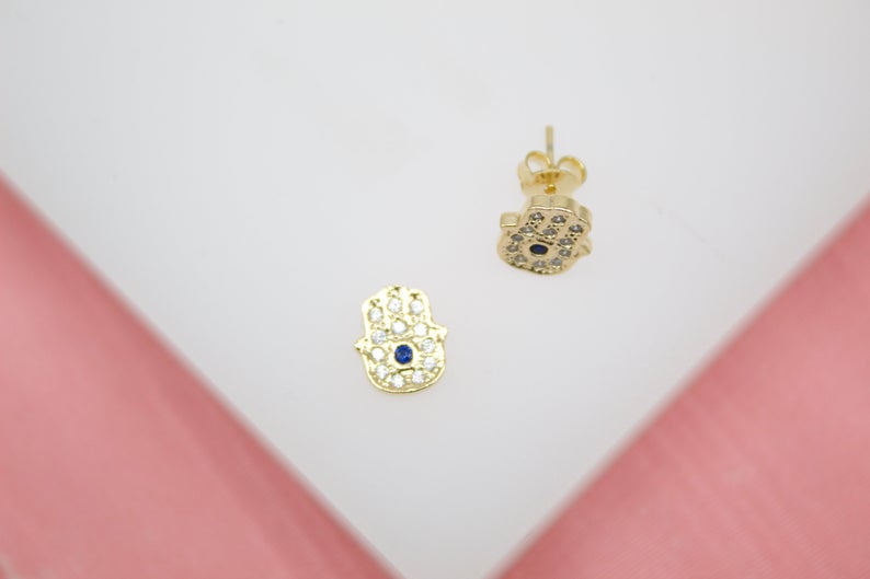 A pair of gold Hamsa hand stud earrings featuring sparkling pave crystals and a central blue stone rest on a white surface.