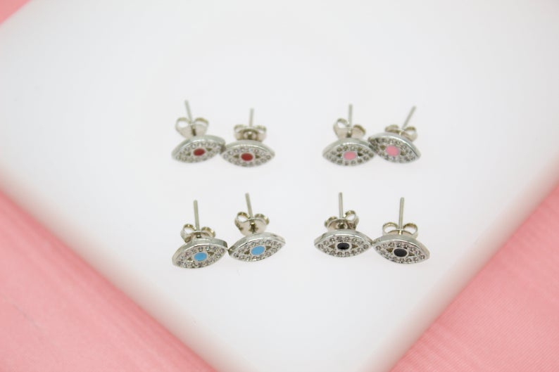 Four pairs of silver evil eye stud earrings with sparkling pavé crystals and colorful enamel centers display on a white surface.