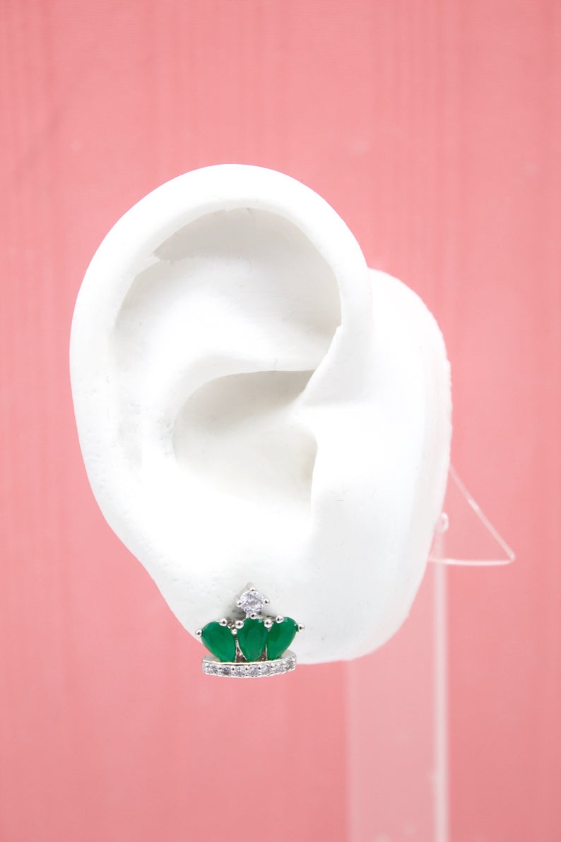 A white model ear displays a crown-shaped silver stud earring featuring three green teardrop stones and sparkling cubic zirconia accents against a soft pink background.