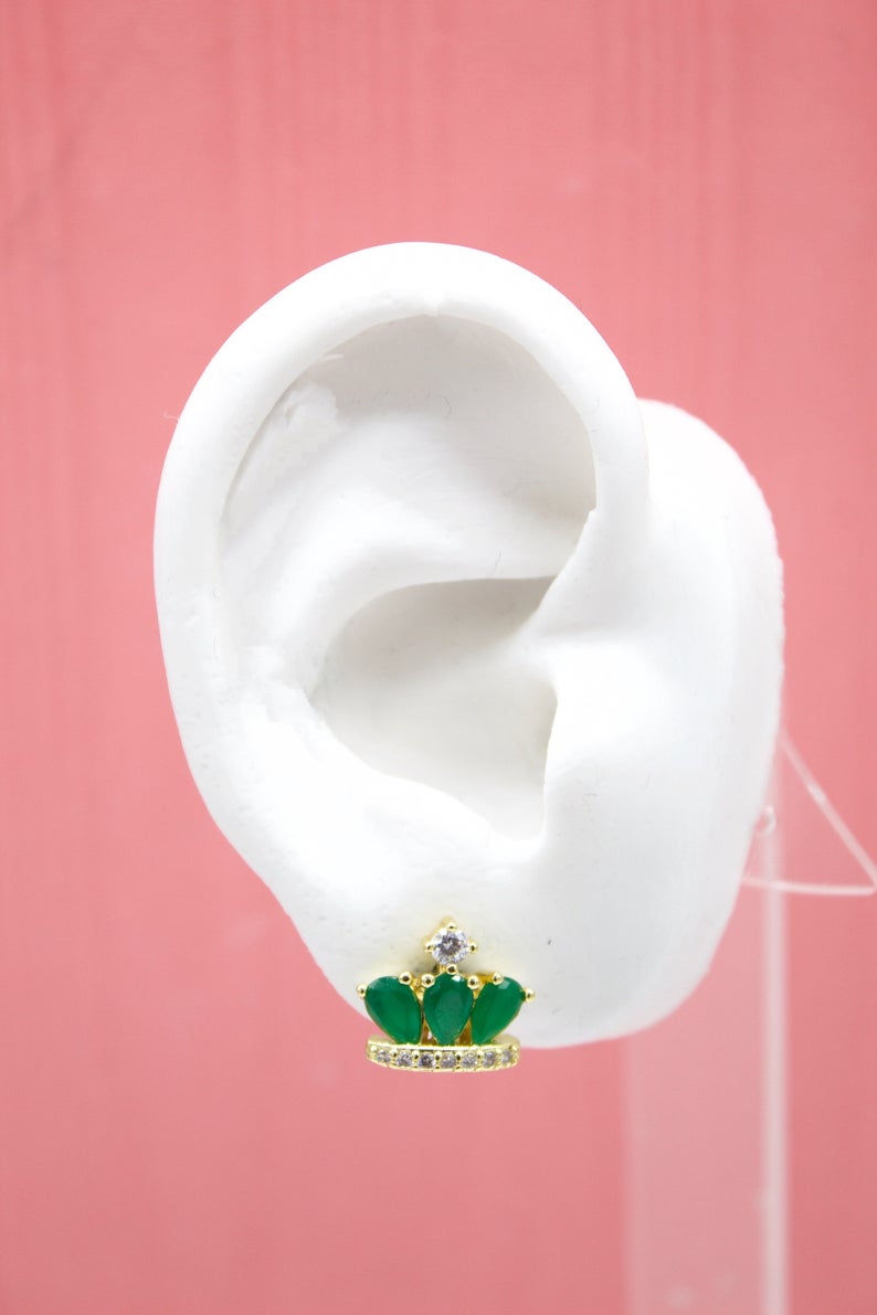 A white mannequin ear displays a gold crown-shaped stud earring adorned with green teardrop stones and sparkling clear crystals.