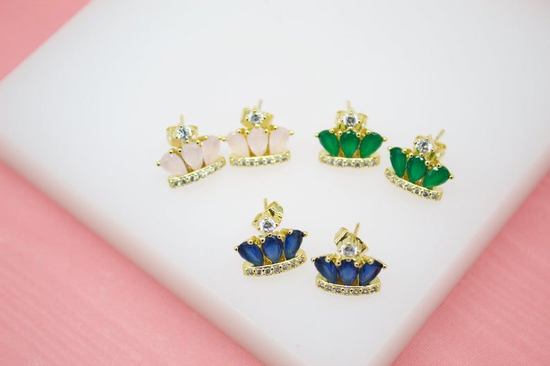 Three pairs of gold crown stud earrings featuring pink, green, and blue teardrop stones rest on a white display surface.