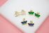 Three pairs of gold crown stud earrings featuring pink, green, and blue teardrop stones rest on a white display surface.