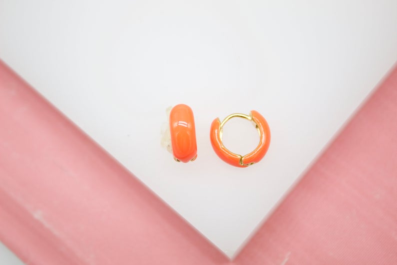 A pair of vibrant neon orange enamel huggie hoop earrings rests on a white display surface against a pink background.