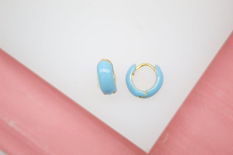 A pair of gold huggie hoop earrings featuring vibrant light blue enamel coating rests on a white and pink display surface.