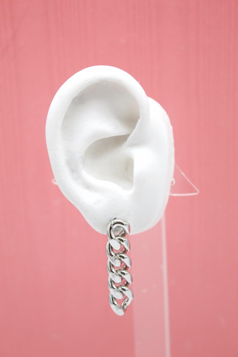 A white mannequin ear displays a stylish silver chunky chain link earring against a solid pink background.