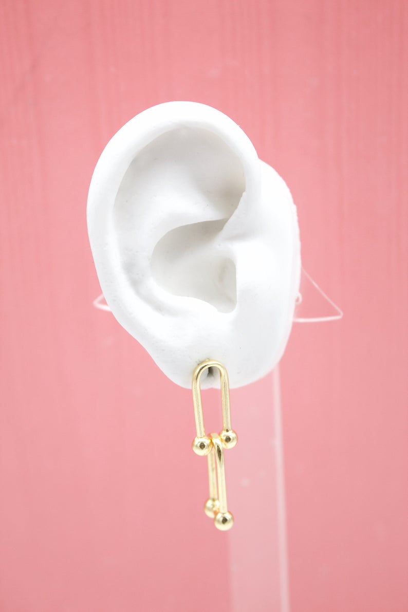 A white mannequin ear displays a modern gold paperclip chain dangle earring against a solid pink background.