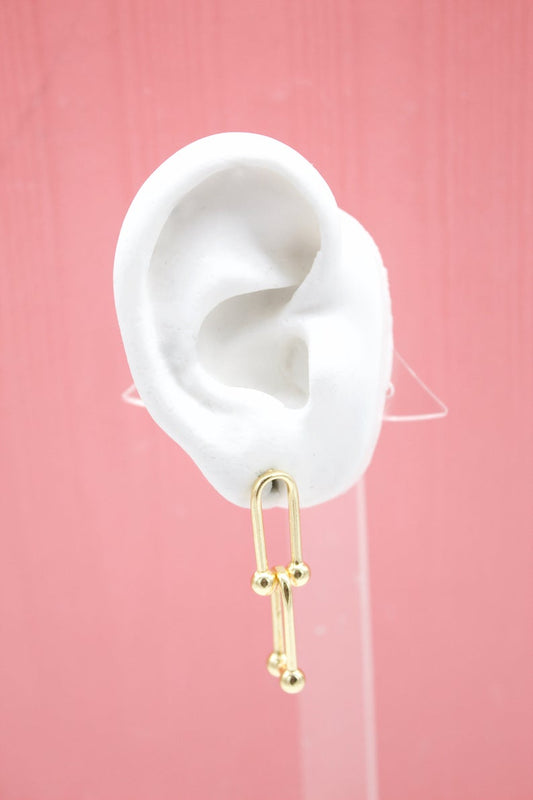 A white mannequin ear displays a modern gold paperclip chain dangle earring against a solid pink background.