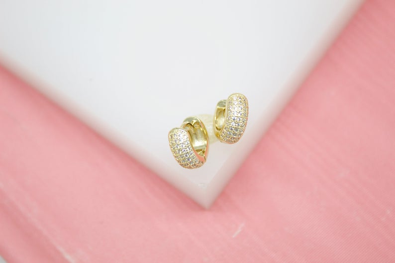 A pair of pavé-set cz gold huggie hoop earrings rests on a white display against a pink background.