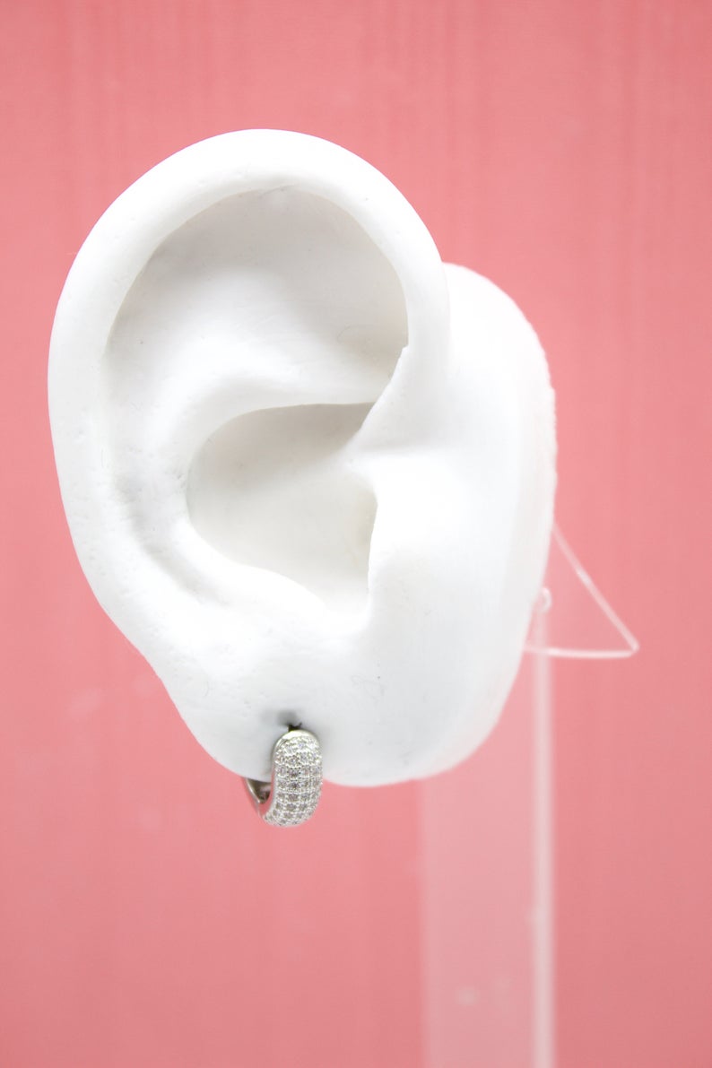 A white ear model displays a sparkling pave hoop earring against a pink background.