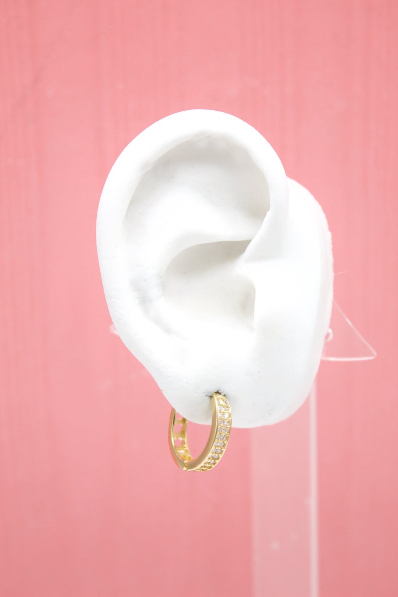 A white mannequin ear displays a sparkling gold huggie hoop earring against a soft pink backdrop.