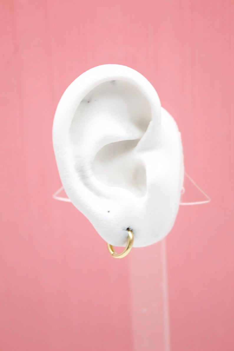 A white mannequin ear displays a small gold hoop earring against a pink background.