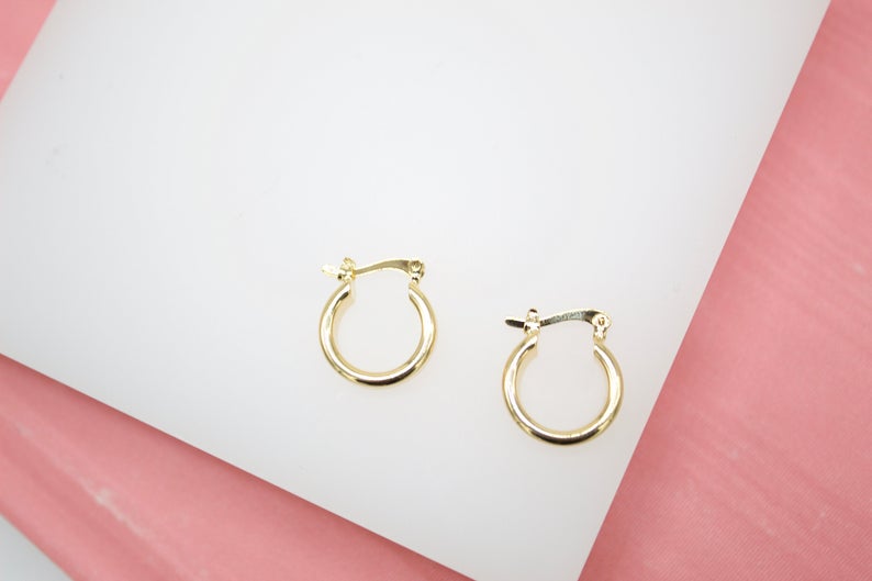 A pair of small gold hoop earrings rests on a white display against a pink background.