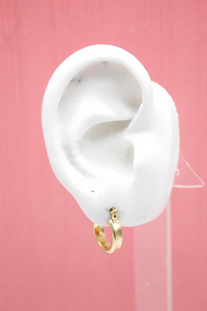 A white mannequin ear displays a small, polished gold huggie hoop earring against a pink background.