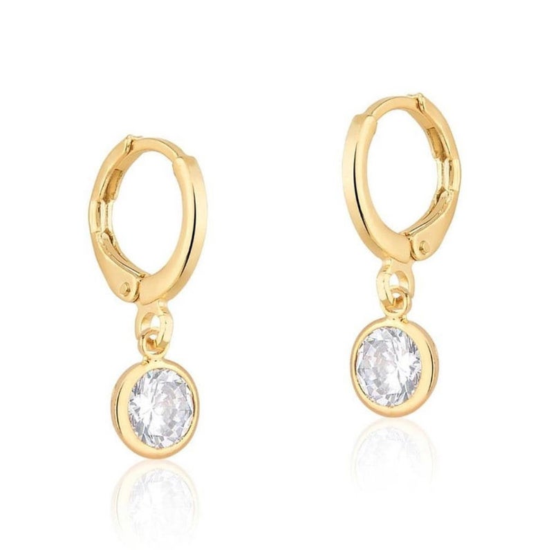 A pair of gold huggie hoop earrings featuring dangling round cubic zirconia stones rests against a clean white background.