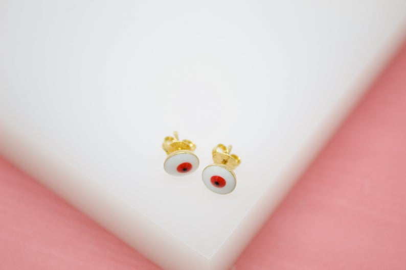 A pair of gold stud earrings featuring round white enamel designs with red and black centers rests on a white surface against a pink background.