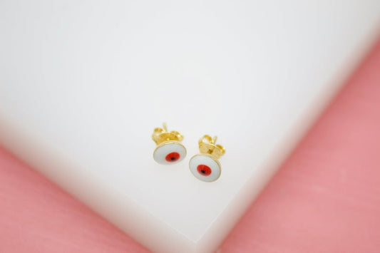 A pair of gold stud earrings featuring round white enamel designs with red and black centers rests on a white surface against a pink background.