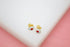 A pair of gold stud earrings featuring round white enamel designs with red and black centers rests on a white surface against a pink background.