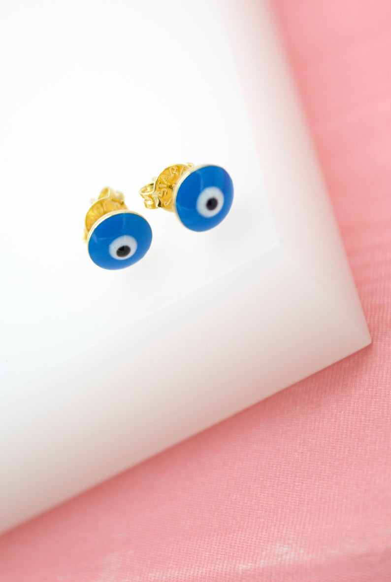 A pair of gold evil eye stud earrings rests on a white display against a pink backdrop.