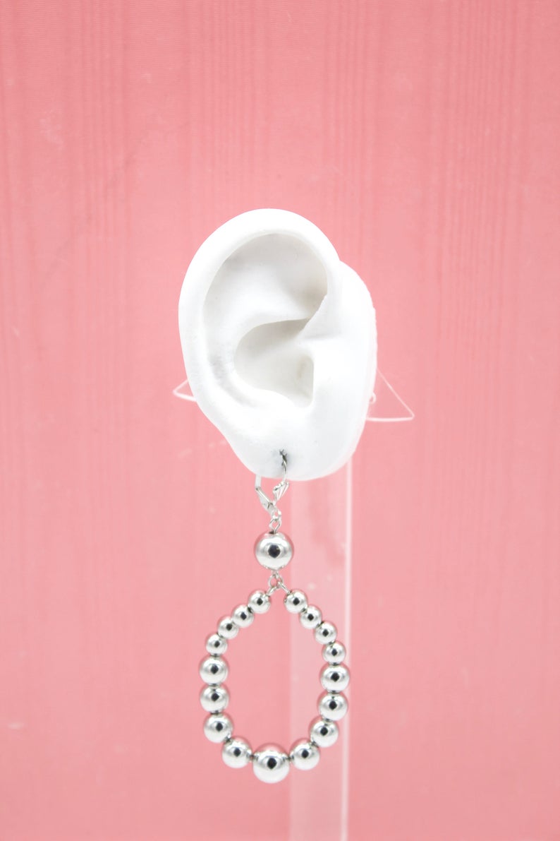 A white mannequin ear displays a silver beaded hoop earring against a pink background.