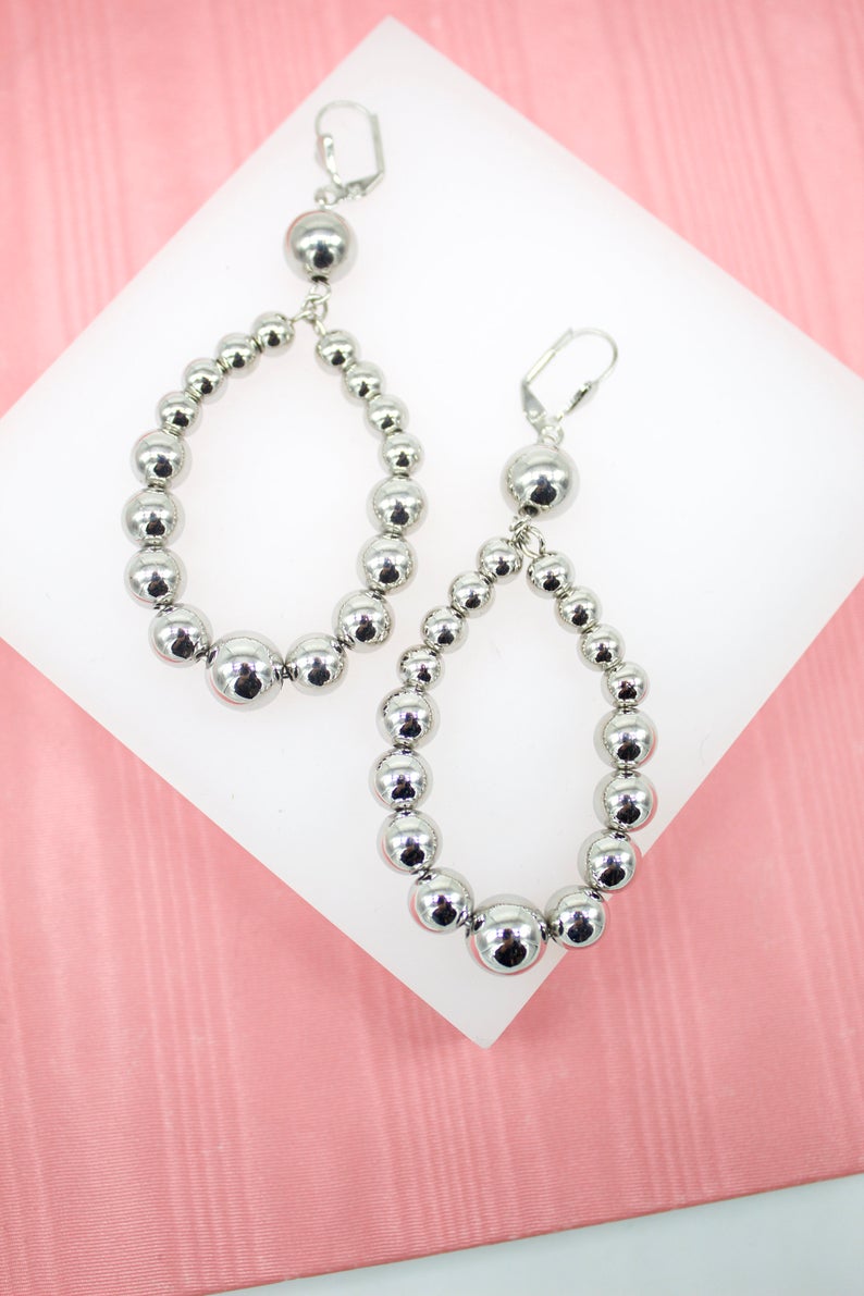 A pair of shiny silver beaded hoop earrings rest on a white display stand against a pink background.