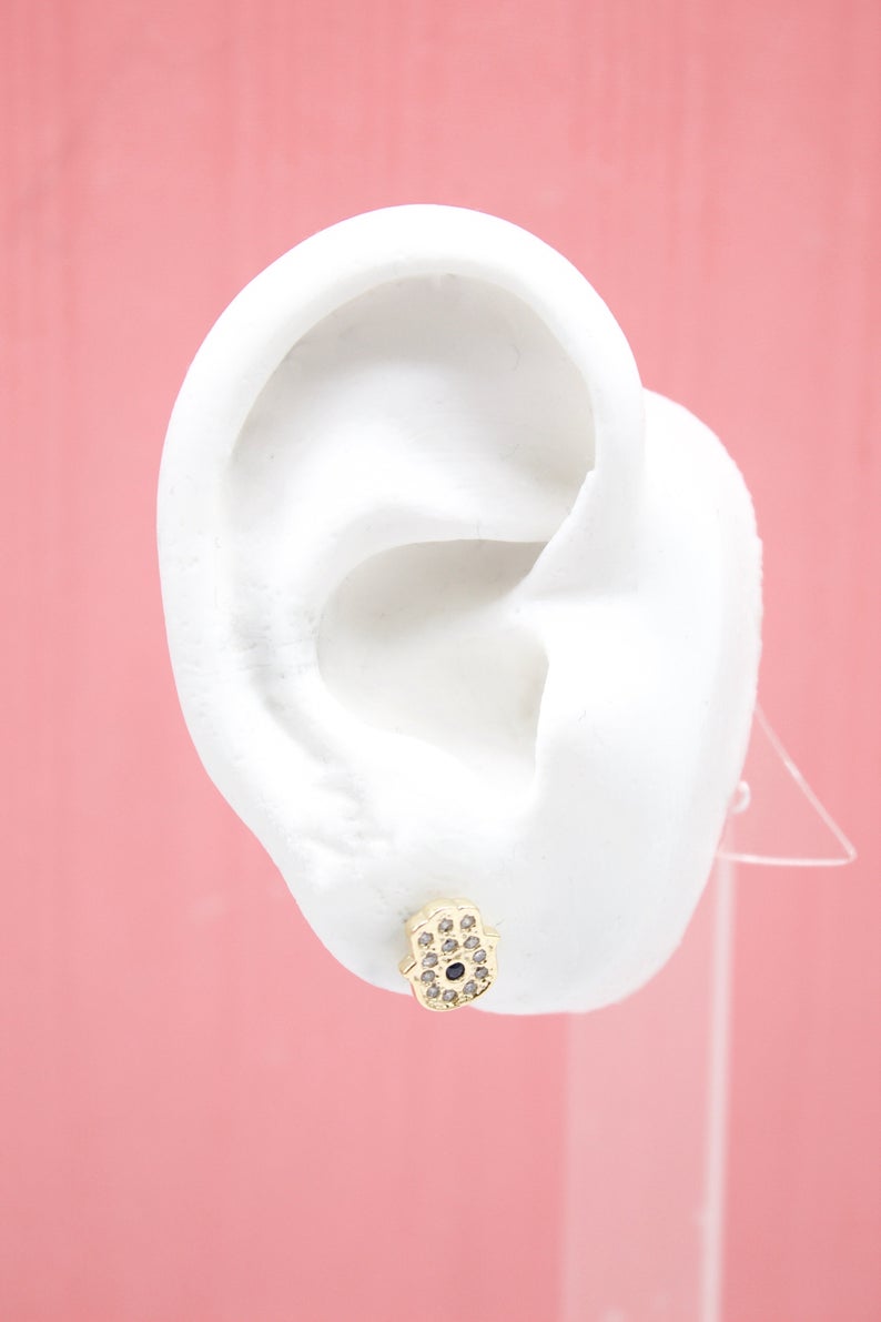 A white mannequin ear displays a gold Hamsa hand stud earring embellished with sparkling clear stones and a dark center stone against a pink background.