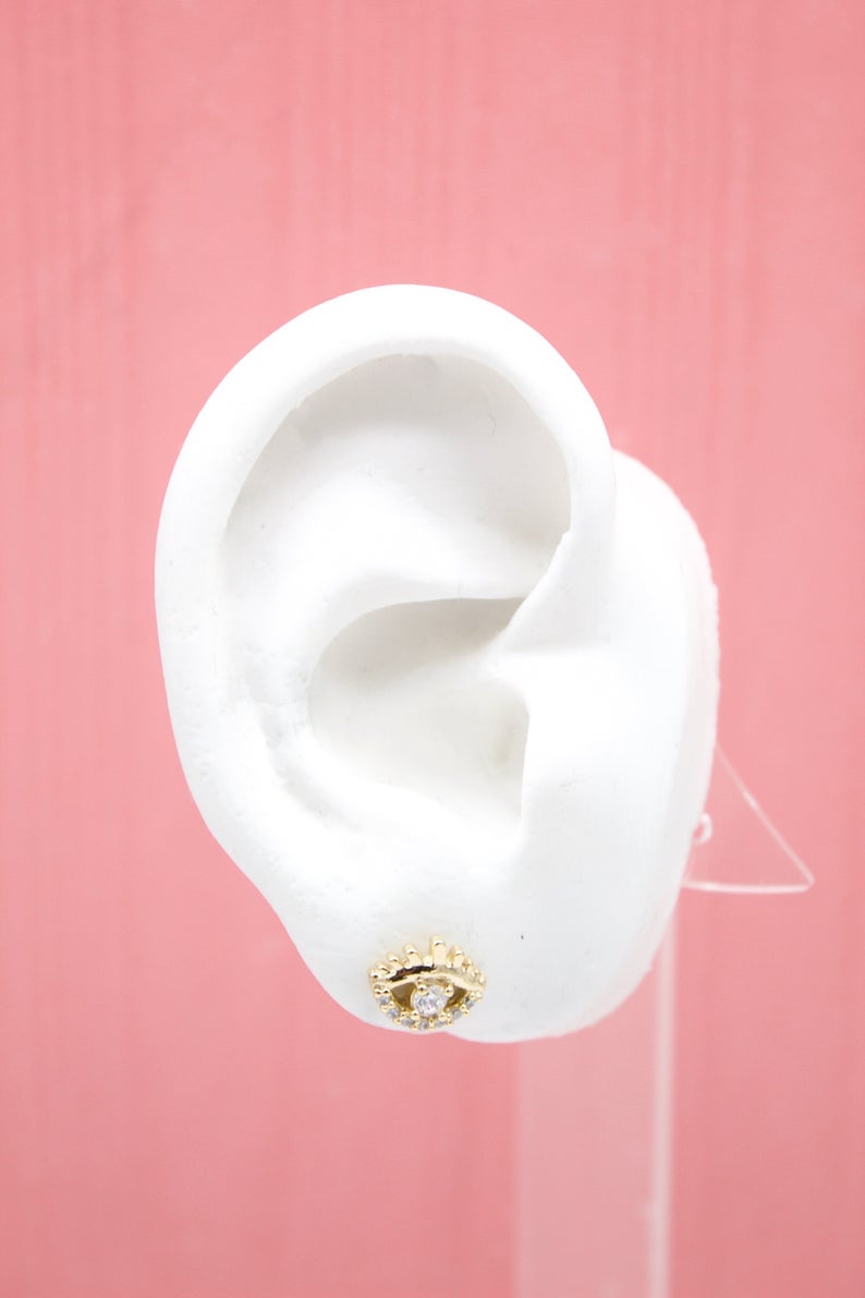 A white mannequin ear displays a gold evil eye stud earring with a central sparkling gemstone against a pink background.