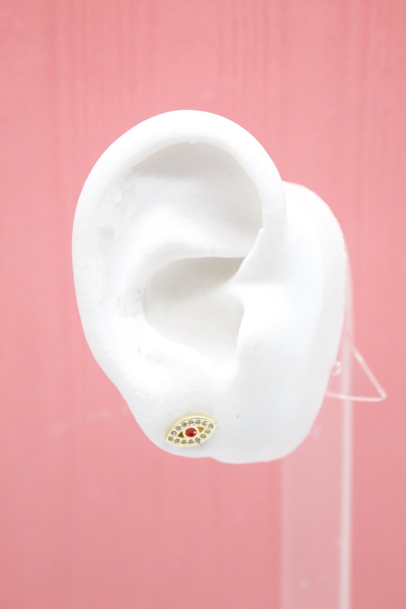 A white mannequin ear displays a gold evil eye stud earring encrusted with sparkling cubic zirconia and a central red stone against a pink background.