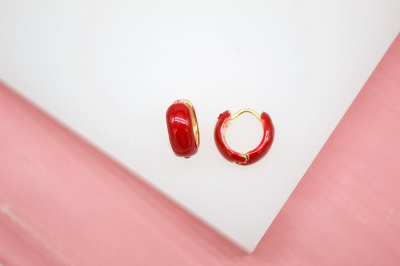 A pair of glossy red enamel gold hoop earrings rest on a white surface against a pink background.