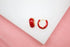 A pair of glossy red enamel gold hoop earrings rest on a white surface against a pink background.