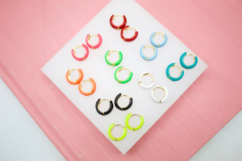 A collection of colorful enamel huggie hoop earrings displays pairs in vibrant shades like red, pink, orange, green, blue, white, black, and yellow arranged neatly on a white display stand against a pink background.