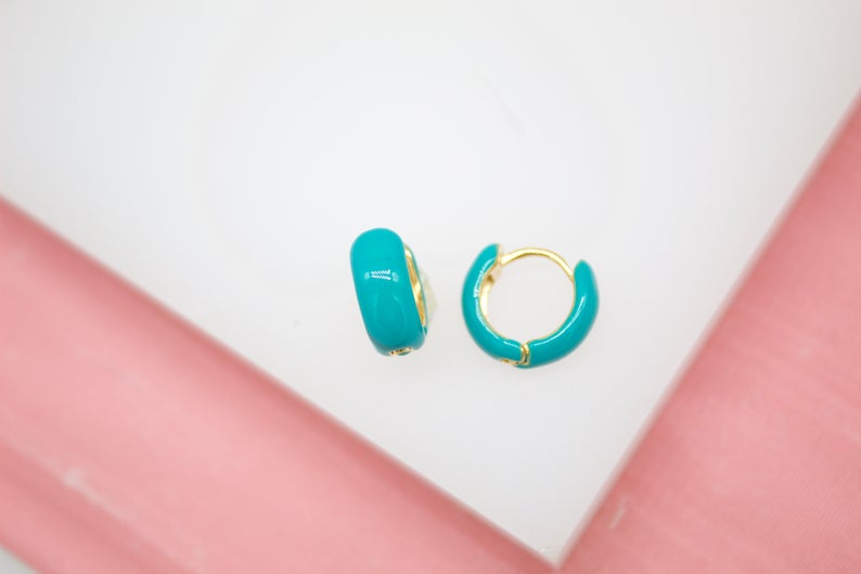 A pair of vibrant turquoise enamel hoop earrings rest on a white display surface against a soft pink background.