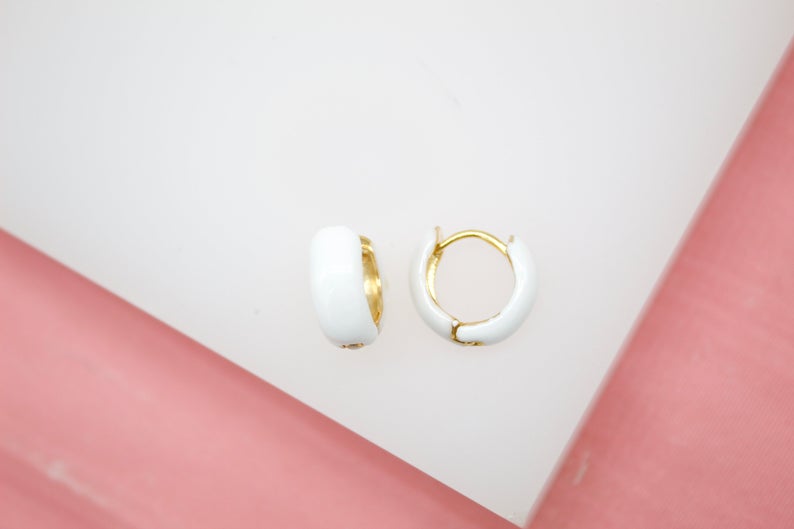 A pair of glossy white enamel gold huggie hoop earrings rests on a white display surface against a soft pink background.