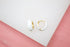 A pair of glossy white enamel gold huggie hoop earrings rests on a white display surface against a soft pink background.