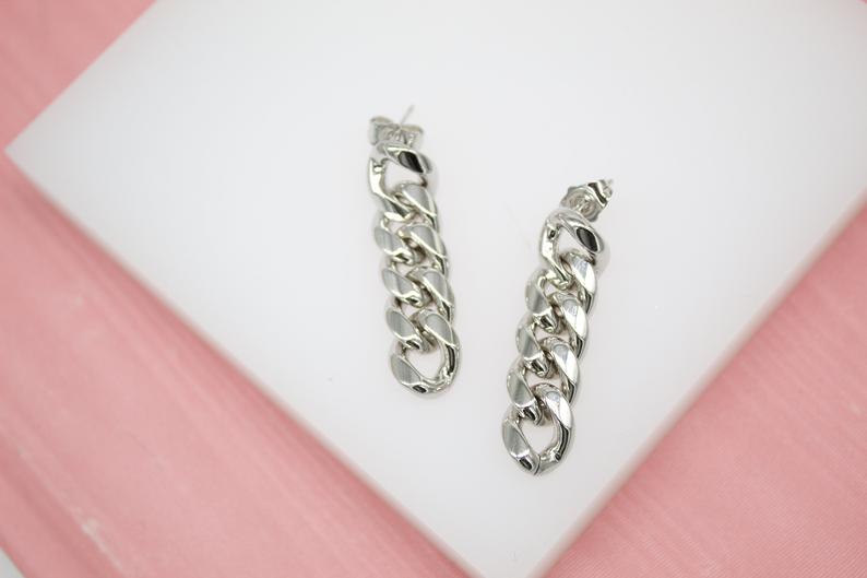 A pair of shiny silver chain link earrings rests neatly on a white display surface against a soft pink background.