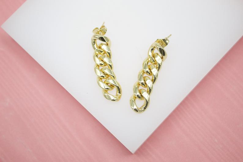 A pair of polished gold cuban link drop earrings rests on a white display surface.