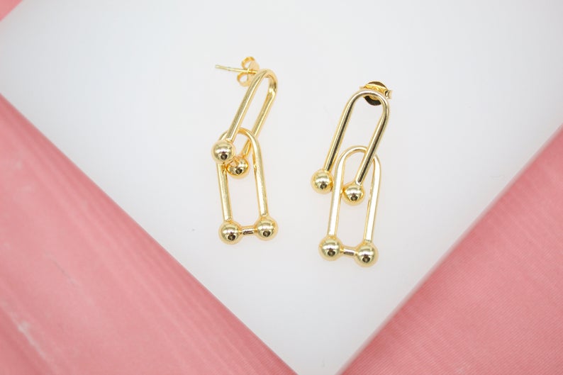 A pair of elegant gold paperclip link earrings with small ball details rests on a white display surface.