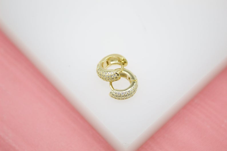 A pair of gold huggie hoop earrings encrusted with sparkling cubic zirconia stones rests on a white display surface against a soft pink background.