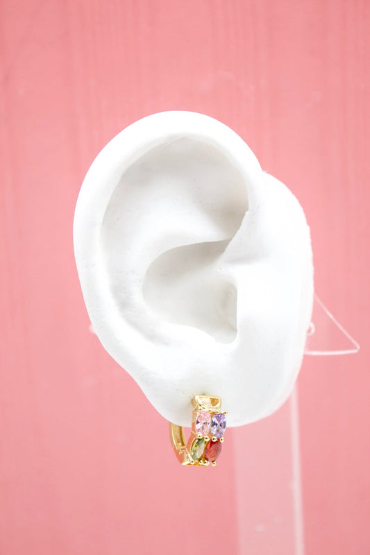 A white mannequin ear displays a gold huggie hoop earring adorned with colorful cubic zirconia stones against a pink background.