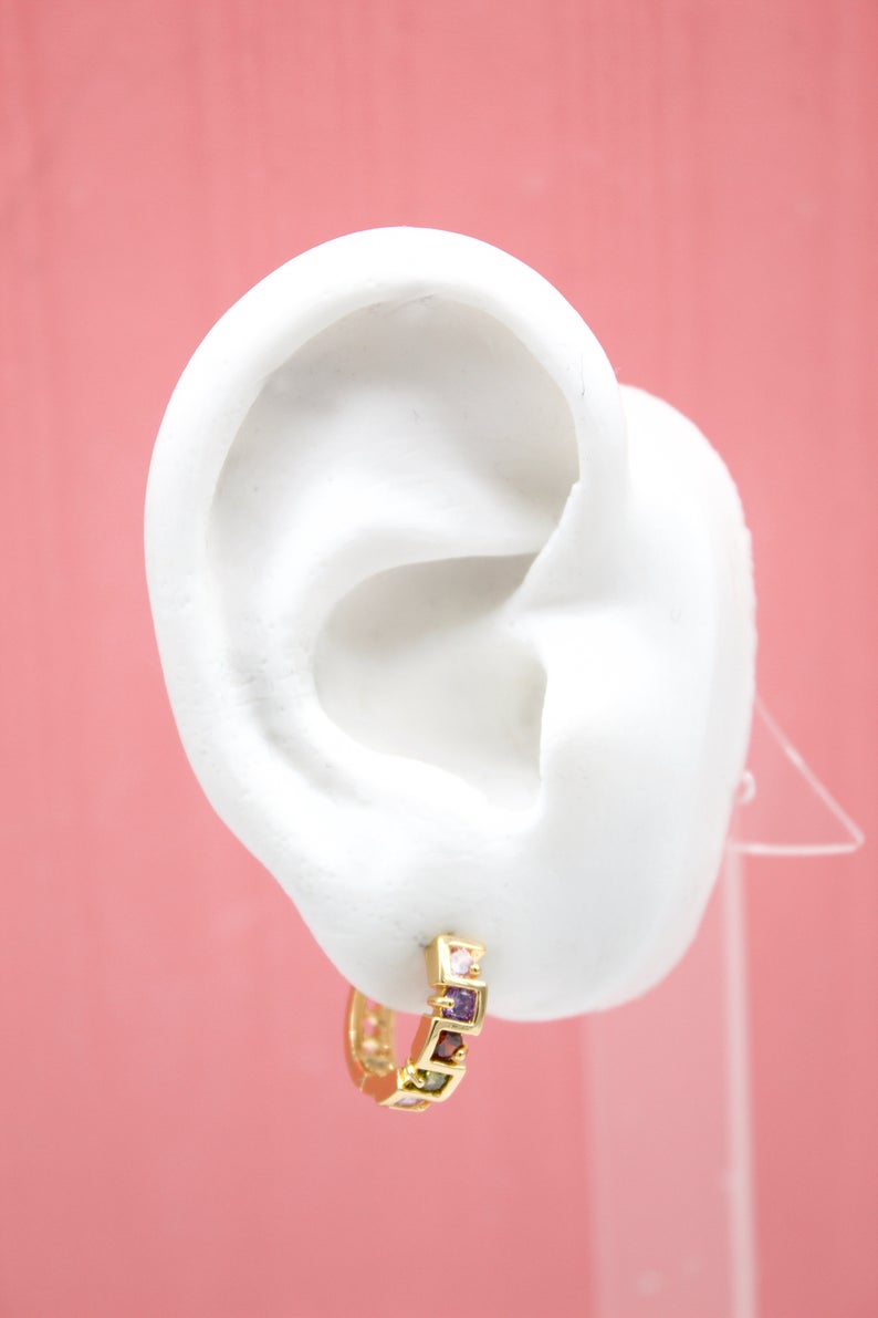 A white mannequin ear displays a gold hoop earring embellished with colorful gemstones against a pink background.