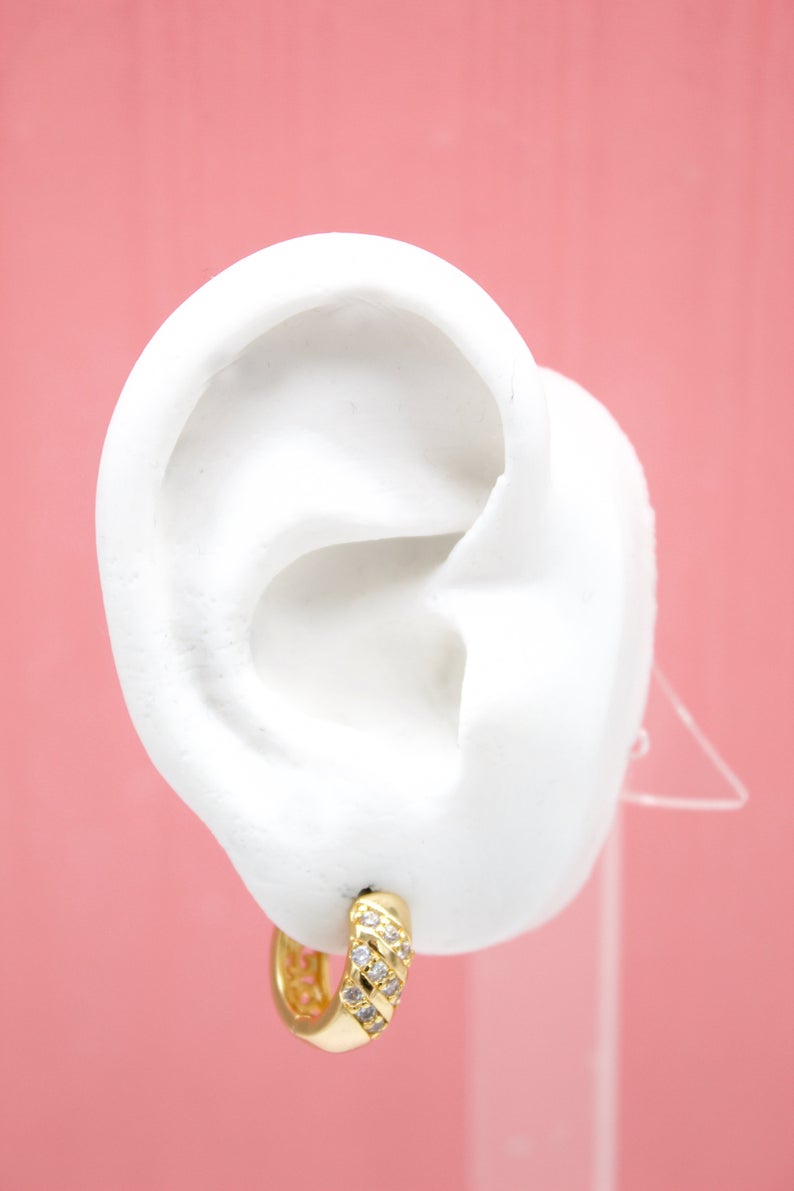 A white mannequin ear displays a sparkling gold hoop earring adorned with small cubic zirconia stones against a pink background.
