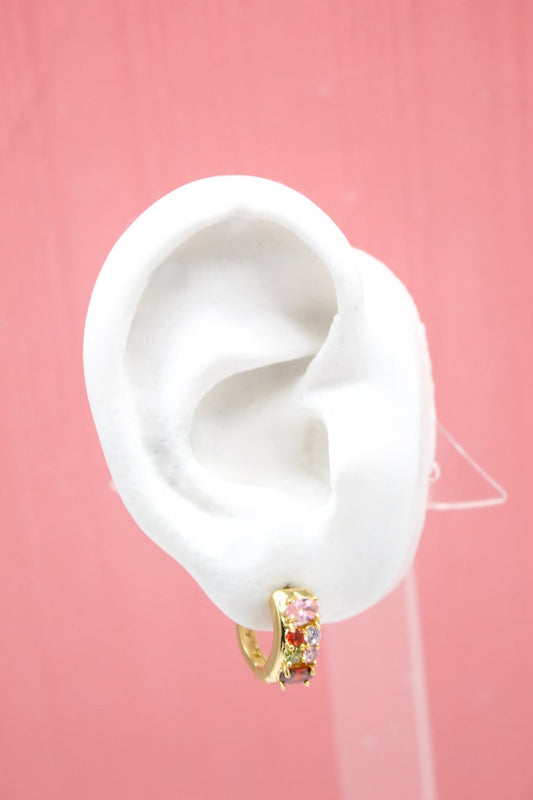 A white mannequin ear displays a gold hoop earring embellished with colorful gemstones against a pink background.