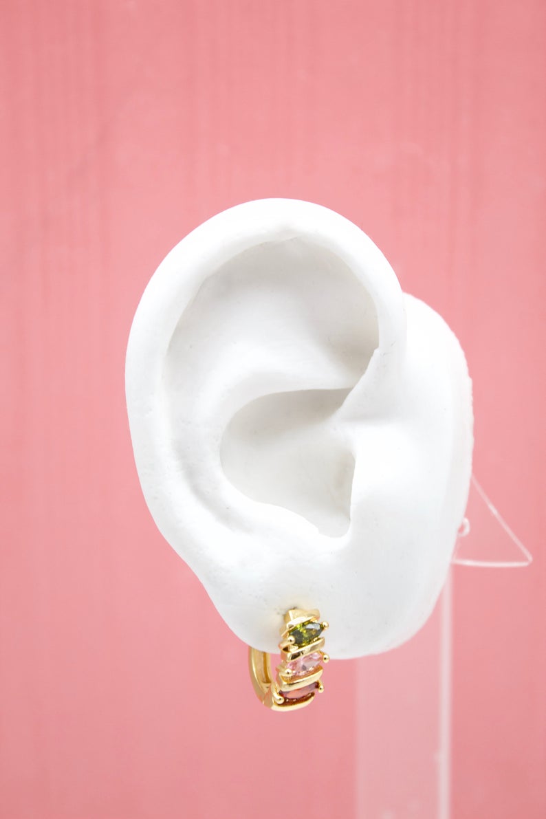 A white mannequin ear displays a gold huggie hoop earring adorned with colorful pink, green, and red gemstones against a soft pink background.