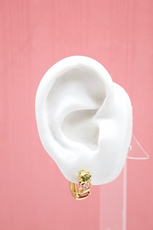 A white mannequin ear displays a gold huggie hoop earring adorned with colorful pink, green, and red gemstones against a soft pink background.