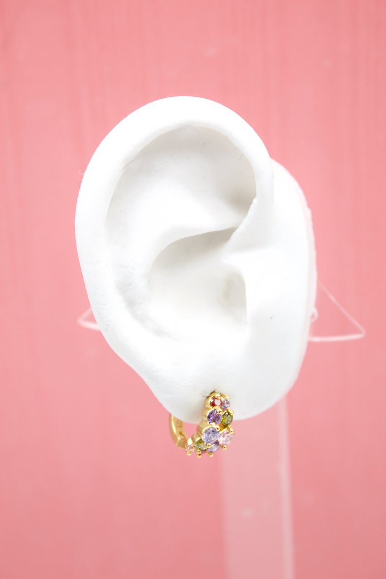 A white mannequin ear displays a gold huggie hoop earring adorned with colorful multicolored cubic zirconia stones.