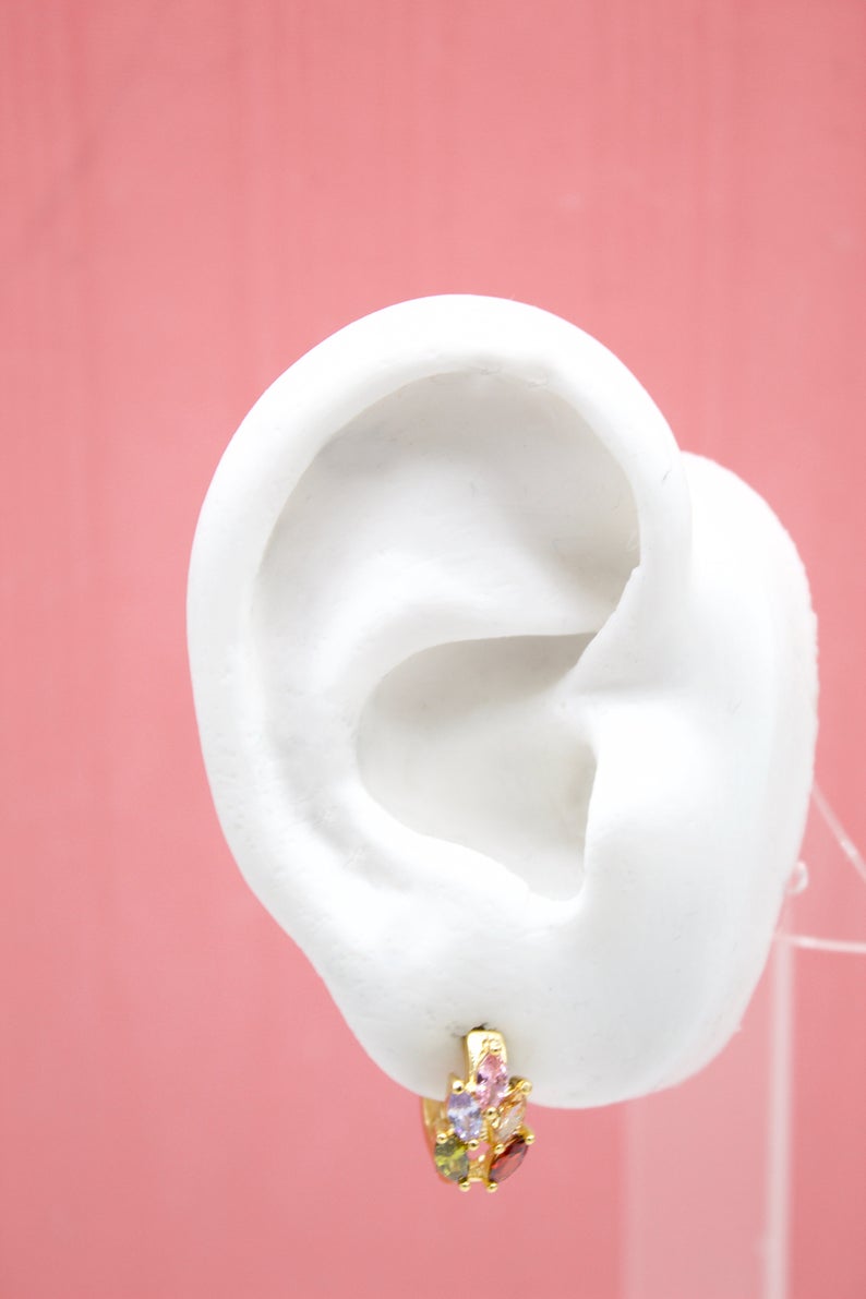 A white mannequin ear displays a gold huggie hoop earring adorned with multicolored gemstone charms against a pink background.
